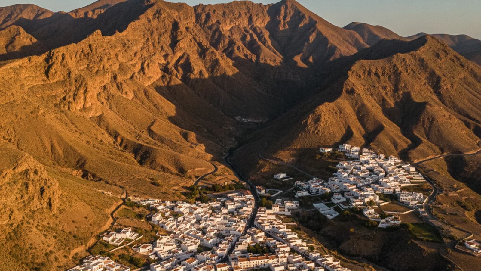 Vista aérea de Betancuria, Fuerteventura, con sus casas blancas agrupadas en el valle y las montañas áridas al fondo, bajo un cielo azul claro.